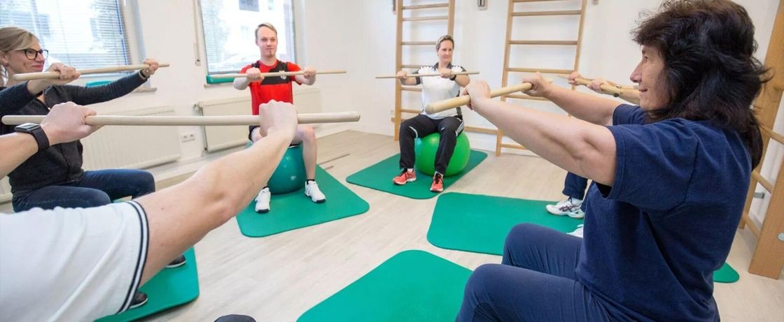 Gruppe von Menschen sitzt auf Gymnastikbällen auf Matten und hält Stöcke mit ausgestreckten Armen nach vorne