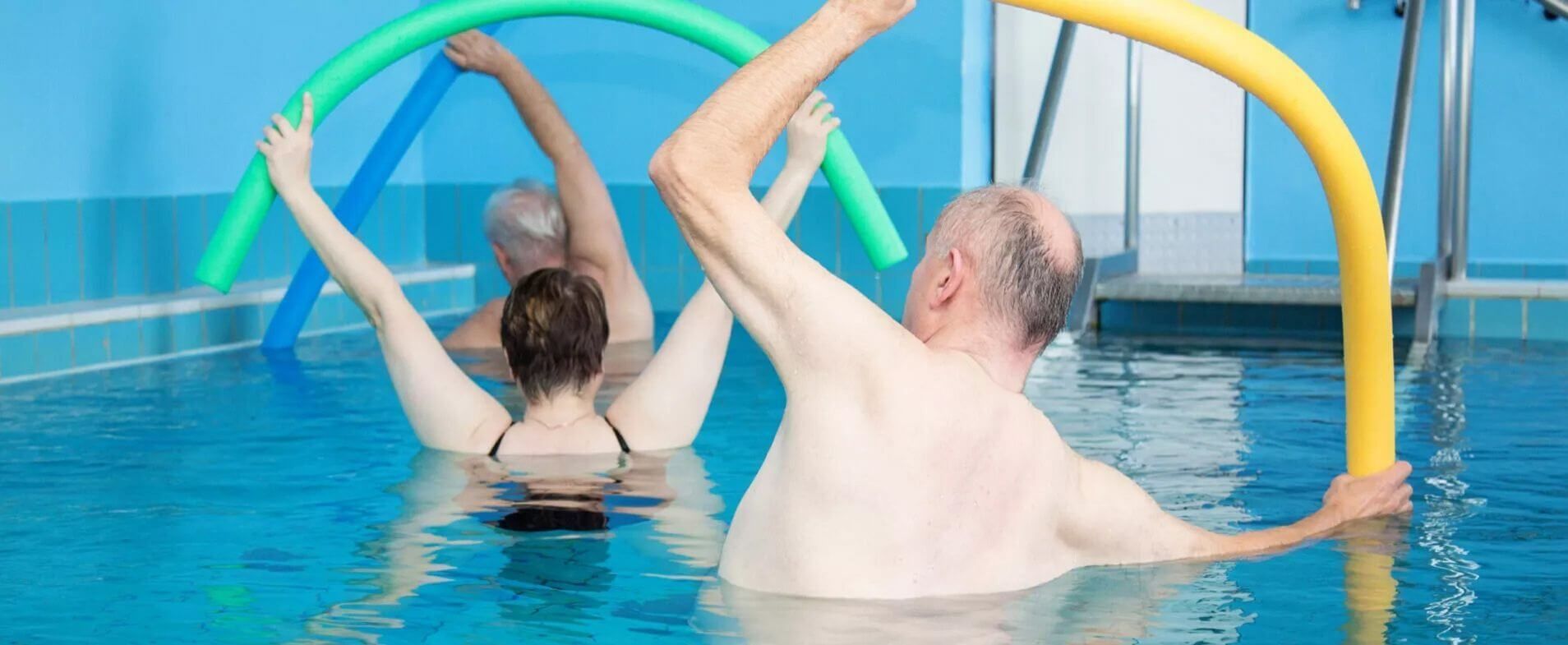 Drei Personen im Schwimmbad halten bunte Schwimmnudeln über dem Kopf und stehen im Wasser